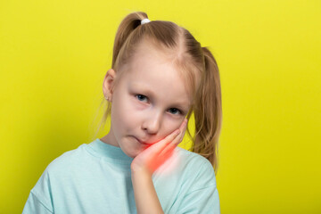 A beautiful little girl of seven years old holds her red, inflamed cheek with her hand. Concept of...