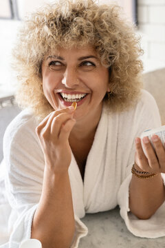 Portrait of a happy, curly-haired woman wearing a white bathrobe and smiling while taking a vitamin capsule. The concept of health, wellness, and daily supplements in a casual home environment.