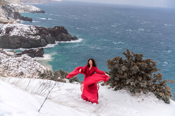 Woman red dress snow sea. Happy woman in a red dress in the snowy mountains by the emerald sea. The wind blows her clothes, posing against sea and snow background.