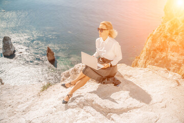 Business woman on nature in white shirt and black skirt. She works with an iPad in the open air with a beautiful view of the sea. The concept of remote work.