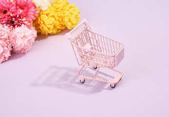 A pink empty shopping cart on a purple background. Multicolored flowers lie on the table.