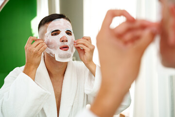 Handsome young man applying sheet mask near mirror in bathroom