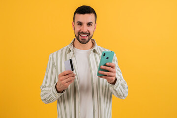 Young adult holding smartphone and showing a credit card smiling at camera on yellow background studio