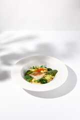 Cod fillet with caviar and zucchini-broccoli garnish, side view on white plate with delicate leaf shadow