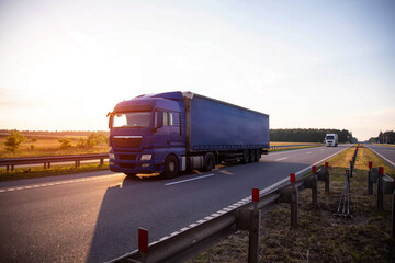 A blue truck with a curtain-sided semi-trailer transports equipment cargo to a factory along a highway against a summer sunset background. Copy space for text