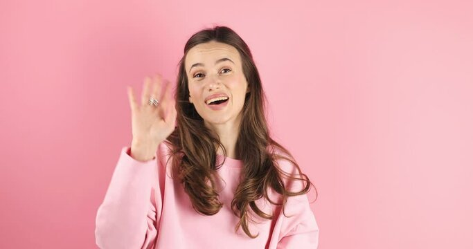 Attractive young woman look by side, turn her head by side to camera and seen somebody, she look happy, smile, waving her hand wear pink long sleeves isolated on pink background. Happy to see you.