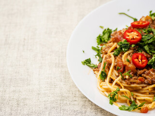 Spaghetti Bolognese dish with pasta, red sauce, minced beef, red peppers and green fresh herbs on white plate and light color table cloth. High quality products in a tasty Italian style dish.