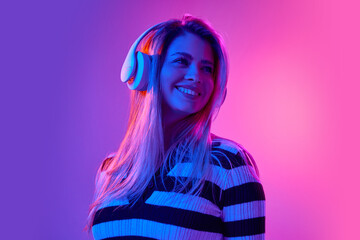 Positive, attractive young woman smiling, wearing headphones and listening to music against pink studio background in neon light. Concept of human emotions, youth, fashion, expression