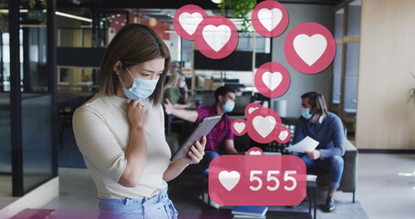 Image of media icons over diverse business people wearing face masks and using tablet