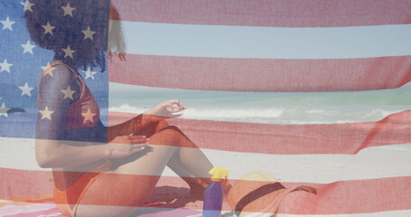 American flag waving against african american woman applying sunscreen at the beach