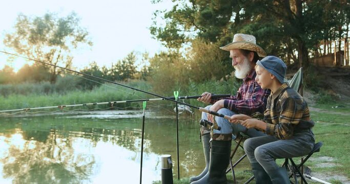 Grandfather man and his small boy grandson throwing rods into pond during joint fishing at sunset. Wonderful moments of family concept.