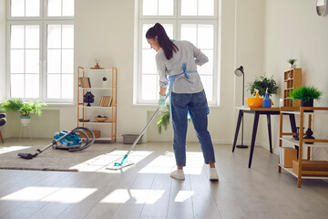 Smiling young woman housewife mopping the floor in the living room at home with detergent. Girl...