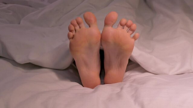 female legs movie and dance toes while lying in bed on a white sheet under a blanket. Happy woman girl with bare legs foot is relaxing and rest on bed in a hotel bedroom in the evening