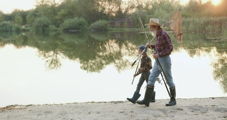 Mature bearded fish man and his small boy grandson together carrying fishing tackles on their shoulders and going fishing on the pond's bank - Powered by Adobe