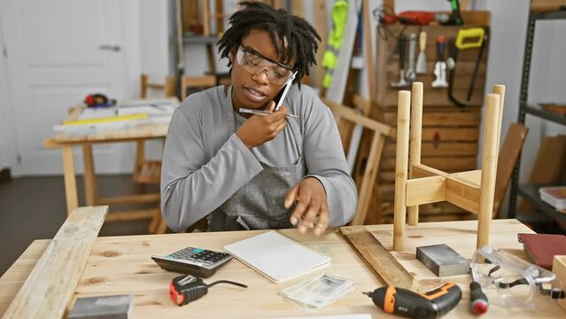 Young african woman with dreadlocks multitasking with phone and danish currency in woodwork studio