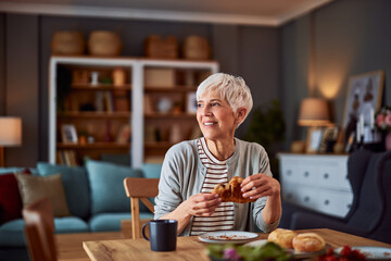 A happy senior adult woman with short hair enjoying a tasty croissant for breakfast while looking...