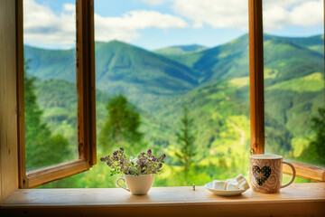 A simple wooden window is wide open and there is a magnificent view of the green mountain summer landscape outside the window. On the windowsill there is a bouquet of flowers, tea and marshmallows. 