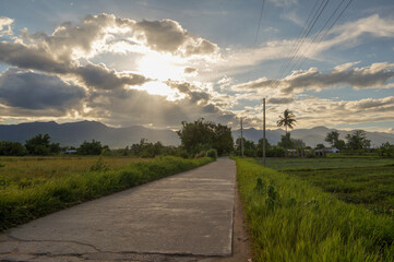 Fototapeta na wymiar The road in the middle of the field in the evening saw clouds blocking the sun and mountains in Thailand. .