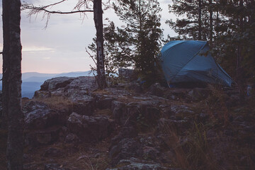 A tent is pitched on a rocky hill in the woods, under an electric blue sky