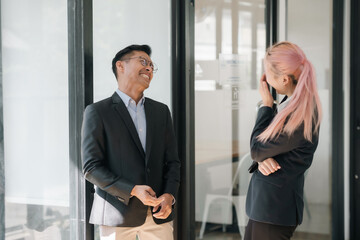 Two asian business professionals laughing during a casual conversation in office corridor. Friendly...