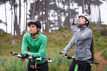 Woman, friends and drinking water in nature with bicycle for cycling, fitness or break on off road trail or path. Thirsty female person or cyclist with beverage in forest or rest for outdoor workout