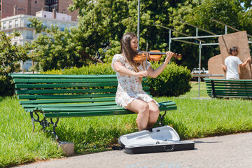 young latin woman busker violinist working sitting on wooden bench with case on floor for money