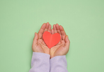 A woman's hand holds a red paper heart on a green background, symbolizing the concept of donation