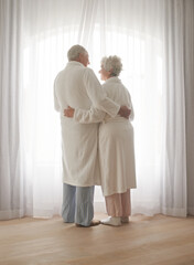 Senior couple, hug and relaxing by window in retirement, love and bonding in pajamas on holiday....