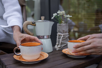 Two hands holding ceramic cups over a wooden table with a coffee maker. Cozy Outdoor Coffee Break with Fresh Brew and Floral Decor