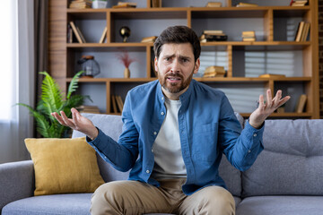 Perplexed young man with beard sitting on sofa, shrugging in confusion