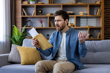 Stressed young man holding envelope with negative emotions on couch at home