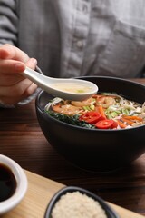 Woman eating delicious ramen with spoon at wooden table, closeup. Noodle soup
