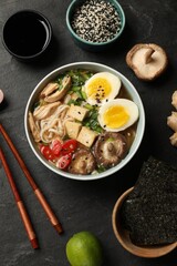 Bowl of delicious ramen, ingredients and chopsticks on black table, flat lay. Noodle soup