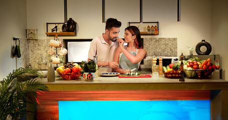 Indian Asian happy smiling young lover couple pair gen z man hug behind woman wife girl indoor home kitchen chop fresh prepare healthy diet salad feed vegan food eat enjoy fun joy spend time