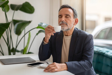 Content senior businessman in a dark blazer savoring a cup of espresso with closed eyes