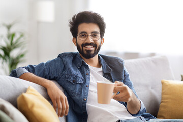 Cheerful young indian man wearing glasses and shirt, enjoying cup of coffee