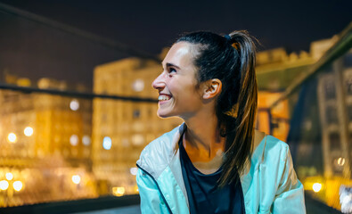 Portrait of cheerful young brunette woman with ponytail looking away in an urban setting at night. Happy female with genuine smile expressing happiness and contentment illuminated by city lights.