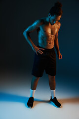 Fototapeta na wymiar young shirtless african american man in black shorts posing in gym on dark lighted background