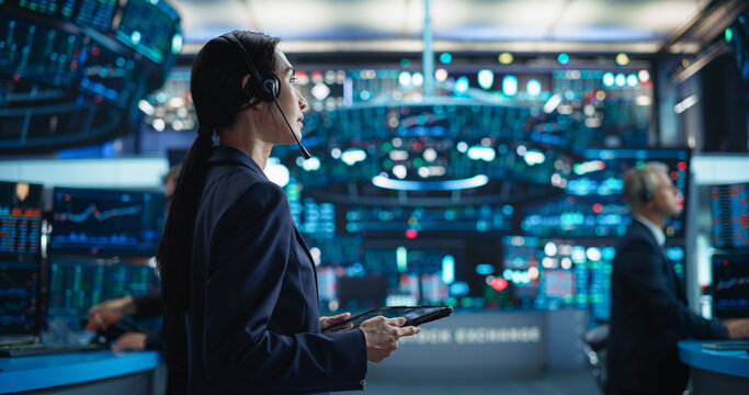 Young Asian Female Broker Working on Tablet Computer on Stock Exchange Floor. Traders Monitoring Real-Time Stocks, Commodities and Exchange Market Data. Professional Investment Agents in Office