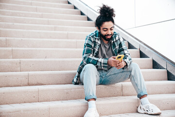 Handsome smiling hipster model. Unshaven Arabian man dressed in summer casual clothes. Fashion male with long curly hairstyle posing in the street. Holds smartphone, uses phone mobile apps