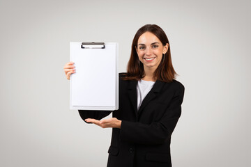 A professional young woman in a black suit confidently presents a blank white clipboard with a smile