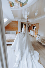 Fototapeta na wymiar Young brunette in long wedding dress and veil in hotel room. A charming bride, full length, in a magnificent white dress on the morning before the wedding.