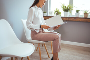 Portrait of confident serious young Asian female sitting on chair in waiting room with electronic tablet, setting her mind up before job interview or meeting with potential business partners