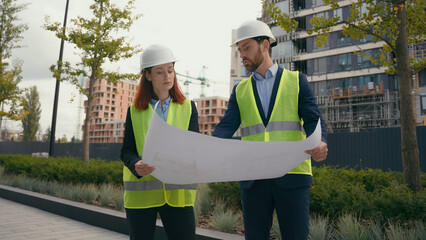 Two people workers builders in safety vests uniform hard hats discuss blueprint document paper plan...
