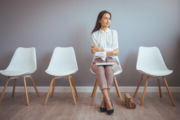 Woman Sitting in a Waiting Room. Serious focused businesswoman holding papers preparing report. Tired of waiting. Businesswoman holding paper and looking away while sitting on chair  - obrazy, fototapety, plakaty