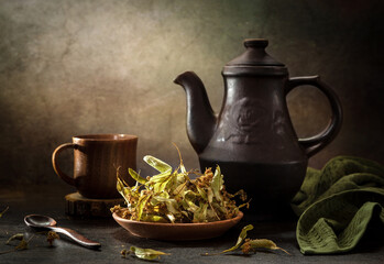 Still life with dried linden blossoms in a plate and linden tea in a teapot on a dark background