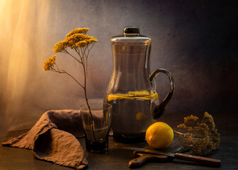 Still life with a glass jug of water with lemon and dried flower on a dark background. A healthy drink