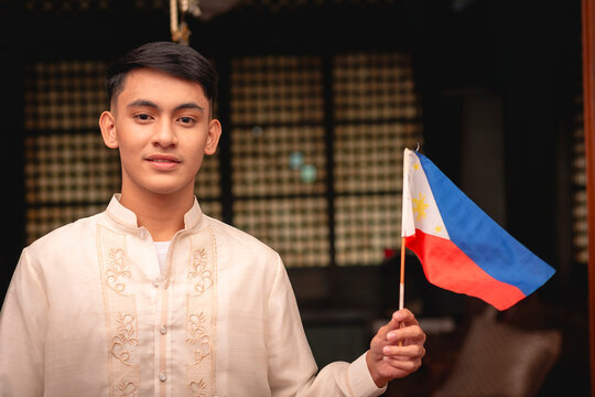 A young FIlipino man in traditional Barong Tagalog shirt and slacks, and holding a Philippine flag, inside an ancestral house.