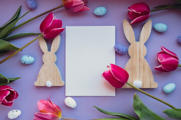 White empty envelope copy space Easter eggs and wooden bunnies with pink tulips on violet pastel background. Greeting card Postcard. Flat lay composition top view. Decoration minimalist modern design
