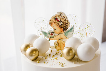 Trendy baptism cake with figure angel with wings and golden decor closeup. Celebration baptism concept. Delicious reception at birthday baby party. Cake on background white photo zone. Side view.